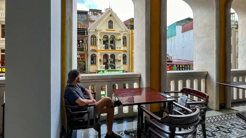 Toti sitting outside Sapa station and enjoying the city | Best areas to stay in Sapa - balcony cafe in Sapa Town facing a classic yellow French colonial-style building with a mountain backdrop.