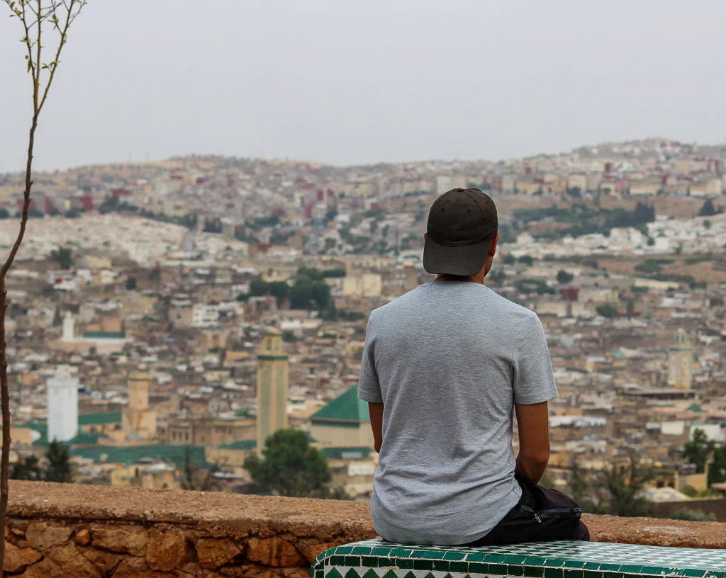 Toti enjoying the view from the iconic Fes Viewpoint - Panoramic landscape of Fes el-Bali rooftops at twilight with the green lights of Kairaouine Mosque minaret visible.