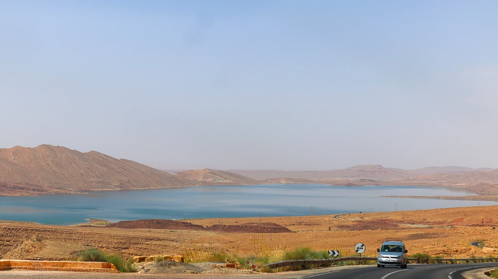 Wide scenic view of the El Mansour Eddahbi Lake near Ouarzazate during a road trip to Merzouga, featuring the contrast of deep blue water against the arid, rugged Moroccan mountain landscape.
